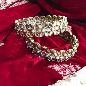 Op de afbeelding: Twee goudkleurige armbanden met een bloemmotief. Elke bloem heeft een spiegelend midden en is versierd met kleine, witte parels. De armbanden liggen op een dieprode fluwelen ondergrond.