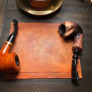 May include: Two tobacco pipes are displayed on a brown leather surface. One pipe features a curved stem, a black mouthpiece, and a light brown bowl. The other pipe has a carved design with a dark, textured bowl and a black stem.