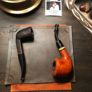 May include: Two tobacco pipes on a brown leather surface. One is dark brown with a curved stem, the other is a lighter orange-brown with a black stem and a light wood band. The pipes are displayed on a brown leather mat.