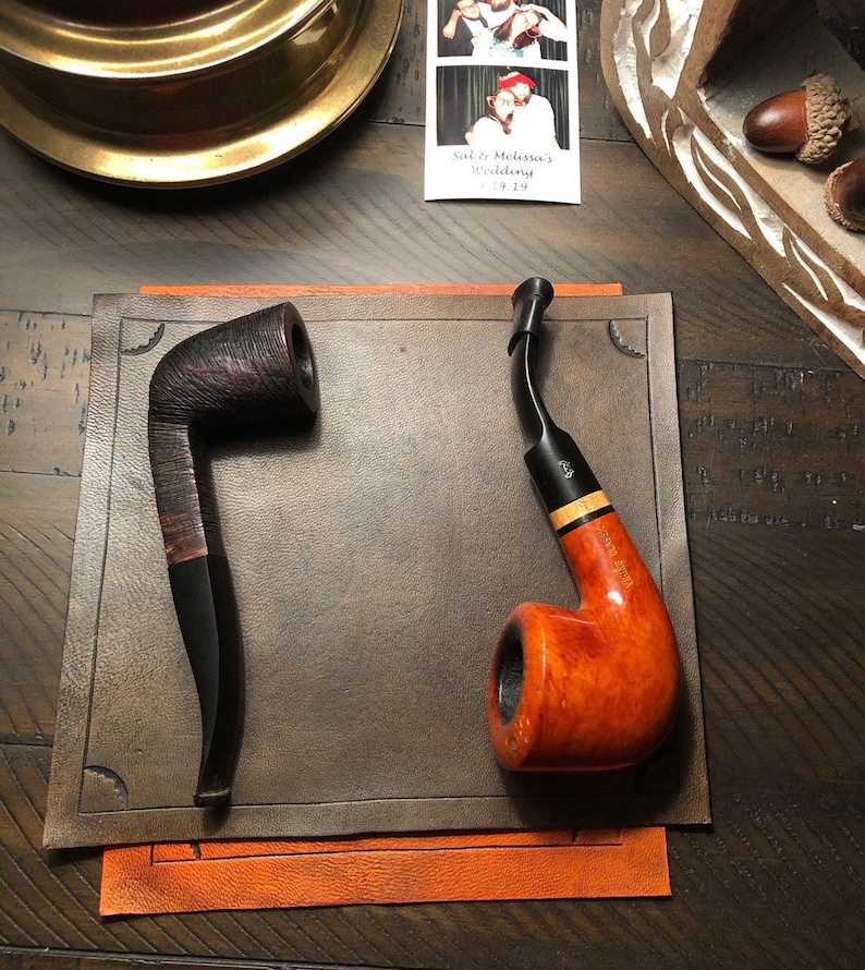 May include: Two tobacco pipes on a brown leather surface. One is dark brown with a curved stem, the other is a lighter orange-brown with a black stem and a light wood band. The pipes are displayed on a brown leather mat.