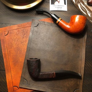 May include: Two tobacco pipes are displayed on textured brown leather placemats. One pipe has a reddish-brown bowl and black stem. The other pipe has a dark, textured bowl and a curved stem. A wedding photo is in the background.