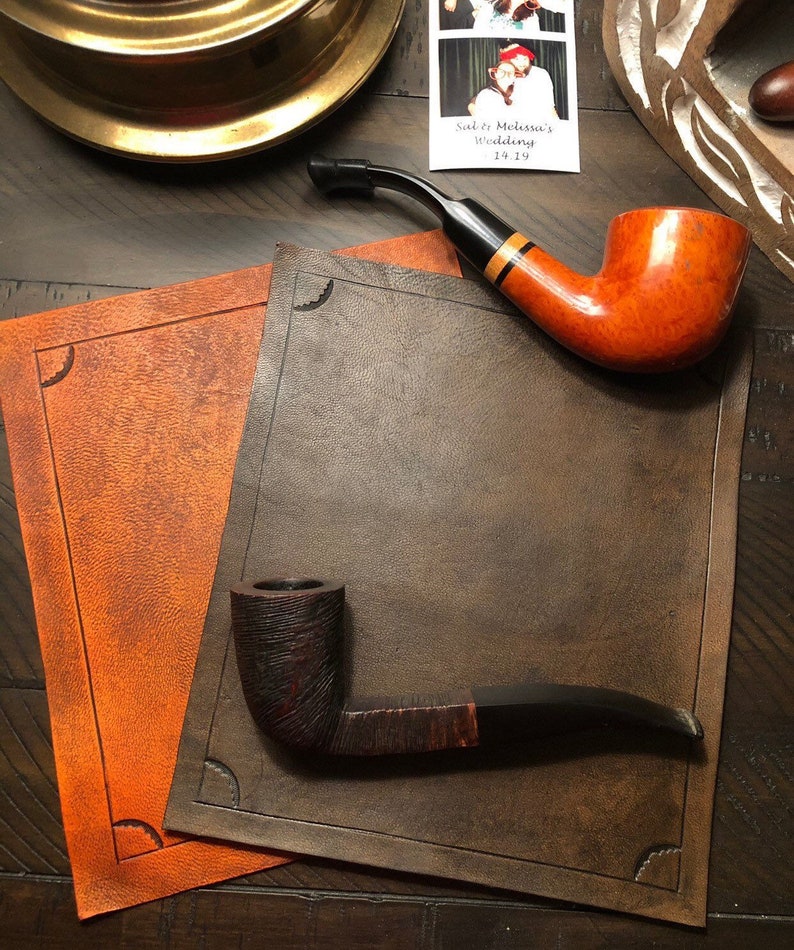 May include: Two tobacco pipes are displayed on textured brown leather placemats. One pipe has a reddish-brown bowl and black stem. The other pipe has a dark, textured bowl and a curved stem. A wedding photo is in the background.