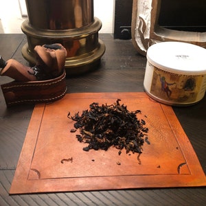 May include: A tobacco pipe with a dark wooden bowl and a black stem, on a brown leather holder. A small pile of loose tobacco sits on a leather mat. A tin with a label is also present.