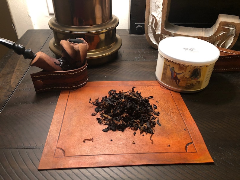 May include: A tobacco pipe with a dark wooden bowl and a black stem, on a brown leather holder. A small pile of loose tobacco sits on a leather mat. A tin with a label is also present.