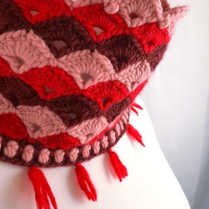 May include: A handmade crochet hat in shades of red, pink, and brown. The hat features a scalloped edge and diamond pattern. Red tassels hang from the bottom edge. The hat is displayed on a white surface.