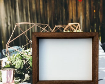 May include: A brown wooden frame with a white mat and a white background. The frame is sitting on a white table with greenery and a gold geometric vase in the background.