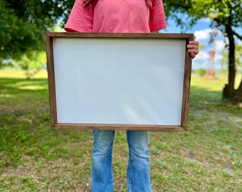 May include: A person is holding a large, rectangular wooden frame with a white canvas insert. The frame is brown with a rustic finish. The person is wearing a pink shirt and blue jeans.