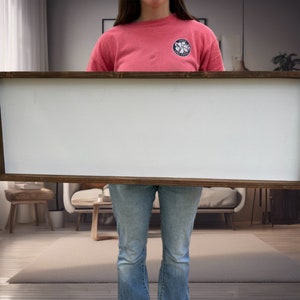 May include: A person holding a large, rectangular, white sign with a brown wooden frame. The sign is blank and has a smooth surface.