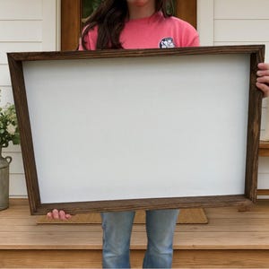 May include: A large, rectangular wooden framed sign with a blank white interior. The frame is a dark brown color. The sign is being held by a person wearing a pink shirt and blue jeans. The background includes a porch and decorative items.