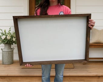 May include: A large, rectangular wooden framed sign with a blank white interior. The frame is a dark brown color. The sign is being held by a person wearing a pink shirt and blue jeans. The background includes a porch and decorative items.