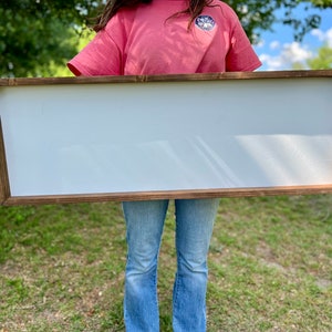 Puede incluir: Un letrero grande, rectangular de madera con una superficie blanca. El letrero est&aacute; siendo sostenido por una persona frente a un campo de hierba.