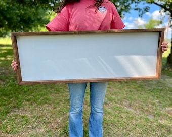 May include: A large, rectangular wooden sign with a white surface. The sign is held by a person in front of a grassy field.