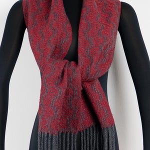 Pu&ograve; includere: Sciarpa tessuta rossa e grigia con bordo a frange. La sciarpa &egrave; drappeggiata su un manichino nero, mostrando il suo intricato motivo e la sua consistenza. La sciarpa &egrave; annodata sul davanti, evidenziando il suo design.