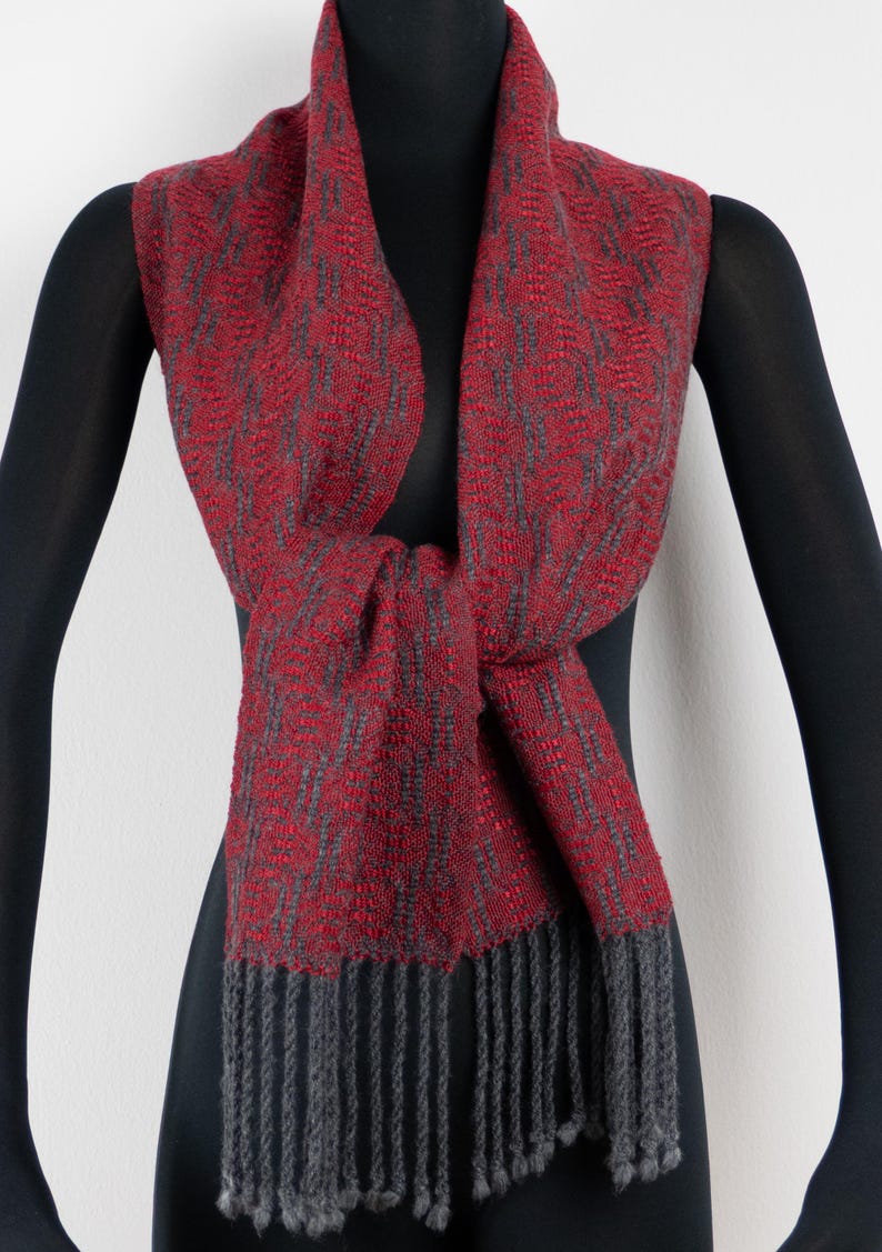 Pu&ograve; includere: Sciarpa tessuta rossa e grigia con bordo a frange. La sciarpa &egrave; drappeggiata su un manichino nero, mostrando il suo intricato motivo e la sua consistenza. La sciarpa &egrave; annodata sul davanti, evidenziando il suo design.