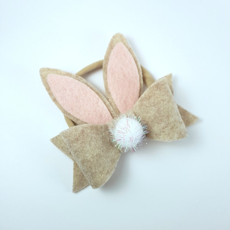 May include: A brown felt bow with pink felt bunny ears and a white pom-pom in the centre. The bow is attached to a brown elastic headband.