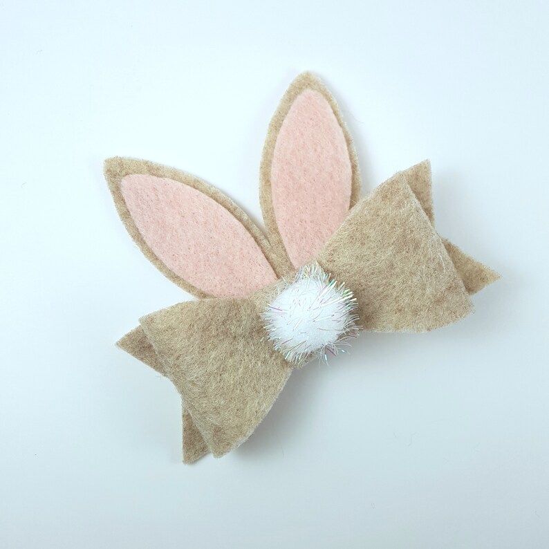 May include: A brown felt bow with pink felt bunny ears and a white pom-pom in the centre.
