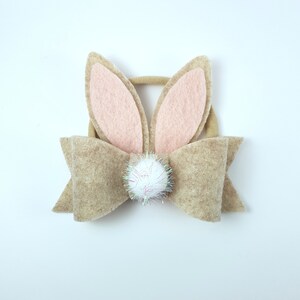 May include: A brown felt bow with a white pom-pom in the centre and pink felt bunny ears attached to the top.