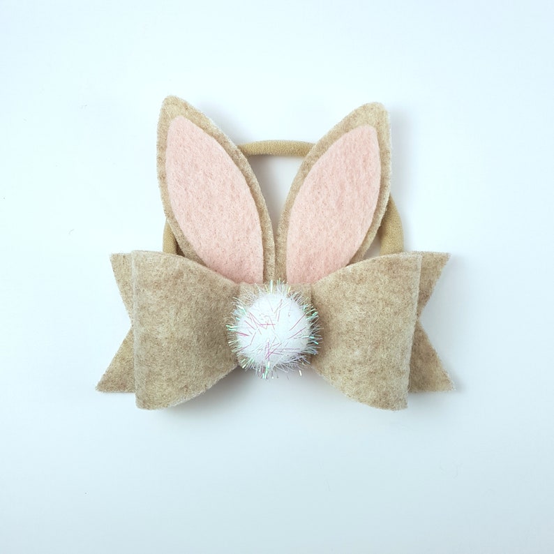 May include: A brown felt bow with a white pom-pom in the centre and pink felt bunny ears attached to the top.