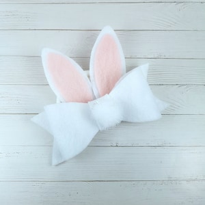 May include: A white felt bow with pink bunny ears. The bow is tied with a knot in the centre and the ears are attached to the back of the bow.