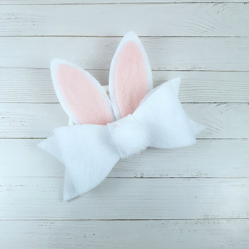 May include: A white felt bow with pink bunny ears. The bow is tied with a knot in the centre and the ears are attached to the back of the bow.