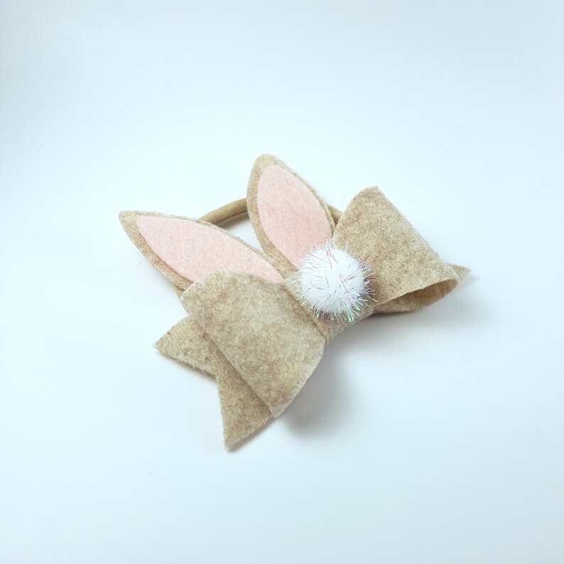 May include: A brown felt bow with bunny ears and a white pom-pom in the centre. The bow is attached to a brown elastic headband.