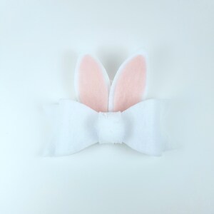 May include: A white felt bow with pink felt bunny ears attached. The bow is tied in a simple knot and the ears are standing up.