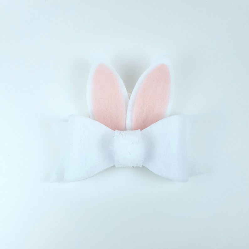 May include: A white felt bow with pink felt bunny ears attached. The bow is tied in a simple knot and the ears are standing up.