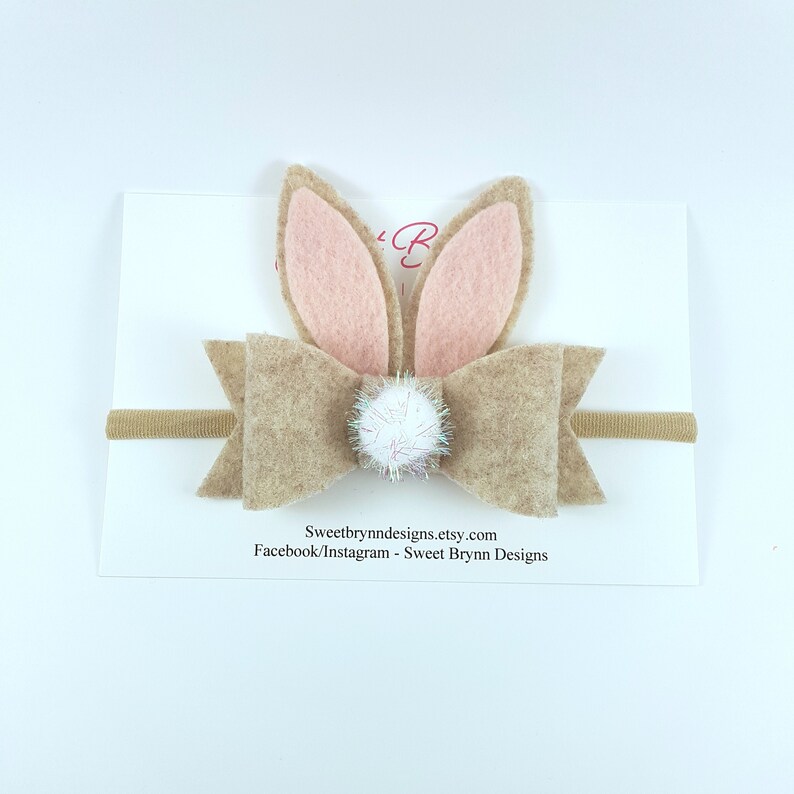 May include: A brown felt bow with pink bunny ears and a white pom-pom accent on a nude elastic headband.