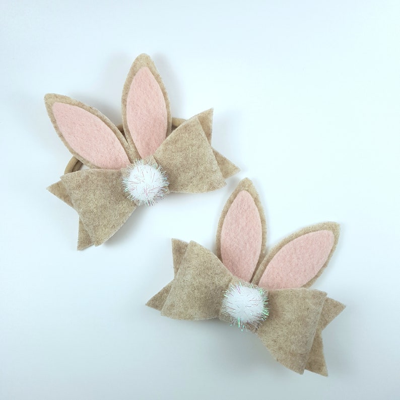 May include: Two brown felt bunny hair bows with pink ears and white pom-poms. The bows are tied with a brown ribbon.
