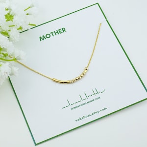 May include: Gold necklace with a delicate chain and a row of small gold beads spelling out the word "MOTHER" in international Morse code. The necklace is displayed on a white card with the word "MOTHER" and the phrase "International Morse Code" printed in green.