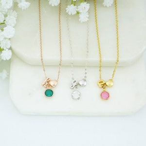 May include: Three gold necklaces with a heart and letter charm and a small round gemstone pendant. The necklaces are on a white surface. The gemstones are green, clear, and pink.