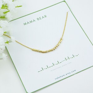 May include: Gold necklace featuring a fine chain and small gold beads spelling out "Mama Bear" in Morse code. The necklace is presented on a white card with the text "Mama Bear" and "International Morse Code" printed on it.