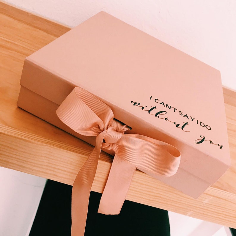 May include: A blush pink gift box with a matching ribbon tied in a bow. The box has the text "I can't say I do without you" printed on the top. The box is sitting on a wooden shelf.