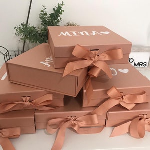 May include: A stack of rose-colored gift boxes with white lettering and heart accents. Each box is tied with a matching satin ribbon. The boxes are arranged in a layered display, showcasing the personalized names on the lids.