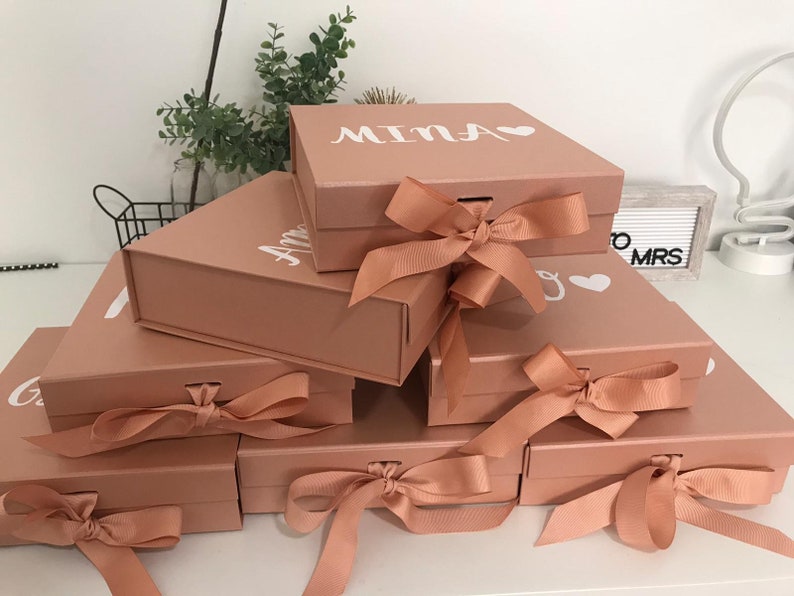 May include: A stack of rose-colored gift boxes with white lettering and heart accents. Each box is tied with a matching satin ribbon. The boxes are arranged in a layered display, showcasing the personalized names on the lids.