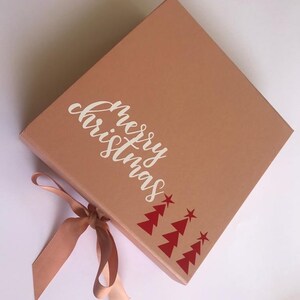 May include: A square, light brown gift box with the words "merry christmas" in white script. Red Christmas trees and stars decorate the corner. A matching ribbon is tied in a bow.
