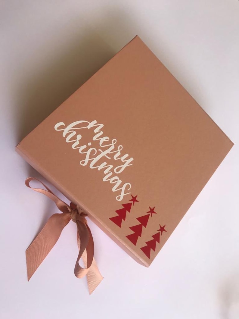 May include: A square, light brown gift box with the words "merry christmas" in white script. Red Christmas trees and stars decorate the corner. A matching ribbon is tied in a bow.