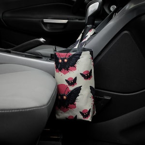 Pode incluir: Um saco de lixo para carro com fundo creme apresentando um padrão repetido de ilustrações de mariposas pretas com detalhes em vermelho. A bolsa é cilíndrica e pendurada na consola do carro.