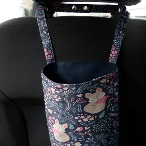 May include: A navy blue car seat organizer with a whimsical fox and floral pattern. The fabric bag hangs from the headrest with adjustable straps. The design features foxes, pink flowers, and green leaves.