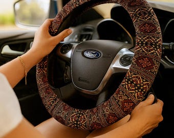 May include: A patterned steering wheel cover in shades of brown, red, and gold. The cover features a detailed geometric design. The steering wheel is black with a silver Ford emblem. The image shows the steering wheel inside a car.