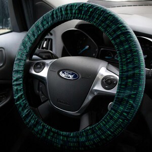 May include: A dark steering wheel cover featuring a repeating pattern of teal binary code digits, specifically ones and zeros, fitted over a black Ford steering wheel with silver accents and integrated control buttons on the spokes.