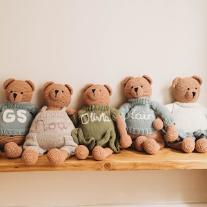 Op de afbeelding: Vijf gehaakte teddyberen die op een houten bank zitten. De beren dragen truien in verschillende kleuren met geborduurde namen: GS, Nora, Olivia, Blair en een witte trui.