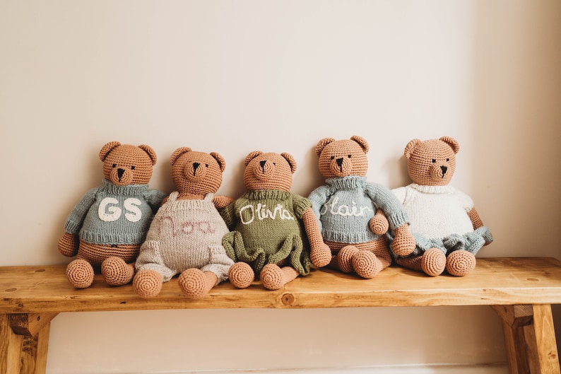 Op de afbeelding: Vijf gehaakte teddyberen die op een houten bank zitten. De beren dragen truien in verschillende kleuren met geborduurde namen: GS, Nora, Olivia, Blair en een witte trui.