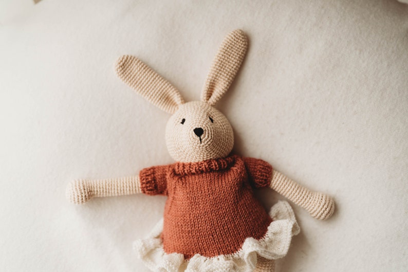 Op de afbeelding: Een gehaakt konijnenknuffel met een roestkleurige trui en een wit kantrokje. Het konijn heeft een beige lichaam en zwarte ogen.