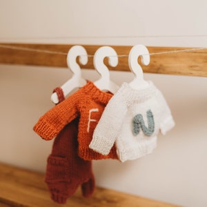 Op de afbeelding: Drie gebreide baby-outfits hangen aan witte hangers op een houten plank. De outfits zijn een rood-oranje trui met bijpassende broek, een witte trui met een blauwe letter "N" op de voorkant en een witte trui met een rode letter "F" op de voorkant.