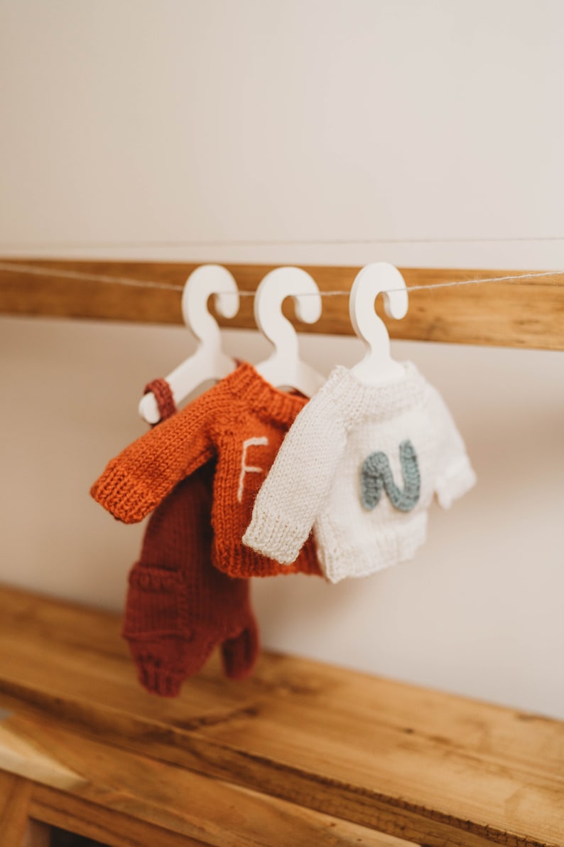 Op de afbeelding: Drie gebreide baby-outfits hangen aan witte hangers op een houten plank. De outfits zijn een rood-oranje trui met bijpassende broek, een witte trui met een blauwe letter "N" op de voorkant en een witte trui met een rode letter "F" op de voorkant.
