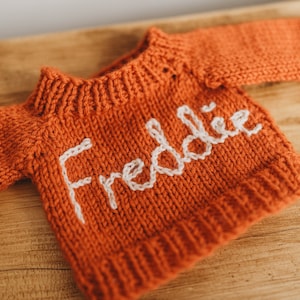 Op de afbeelding: Een kleine, oranje gebreide trui met de naam "Freddie" geborduurd in wit op de voorkant.