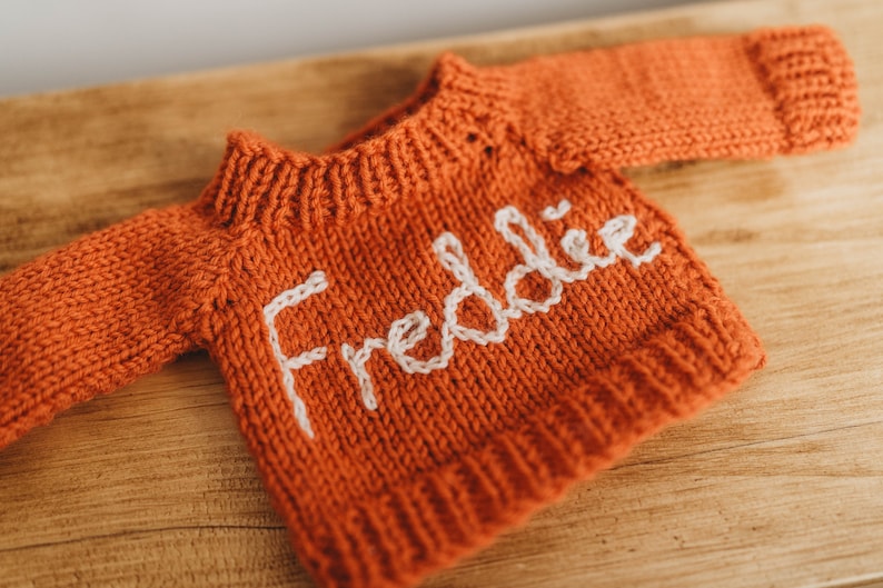 Op de afbeelding: Een kleine, oranje gebreide trui met de naam "Freddie" geborduurd in wit op de voorkant.