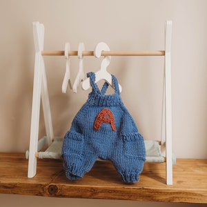 Op de afbeelding: Een witte houten poppenkleerhanger met een blauwe gebreide baby-romper die aan een witte hanger hangt. De romper heeft een rode viltletter 'A' op de voorkant.
