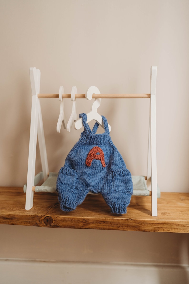 Op de afbeelding: Een witte houten poppenkleerhanger met een blauwe gebreide baby-romper die aan een witte hanger hangt. De romper heeft een rode viltletter 'A' op de voorkant.